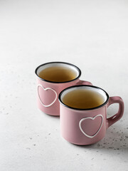 Two pink cups with hearts with tea standing on a white textured background. Drinking beverages together on St. Valentine's day