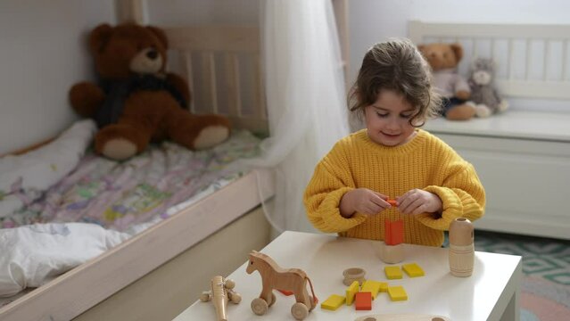 Happy and relaxed little girl with curly hair enjoys playing building a tower with yellow wooden blocks eco-friendly toys in her room.Child development.