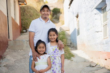 Latino father with his two happy daughters outside his house in rural area-Hispanic father hugging...