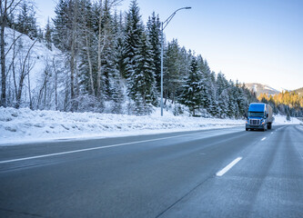 Snowy and icy winter highway mountain road with driving on it industrial big rig blue semi truck with semi trailer in Montana