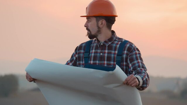 Smiling builder architect reading draft in field. Man examining blueprint while standing on field in countryside. Look around. At sunset. Slow motion.