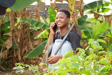 An African female farmer, business woman or entrepreneur happily making a call while standing on a farmland with a farming hoe on her shoulder - obrazy, fototapety, plakaty