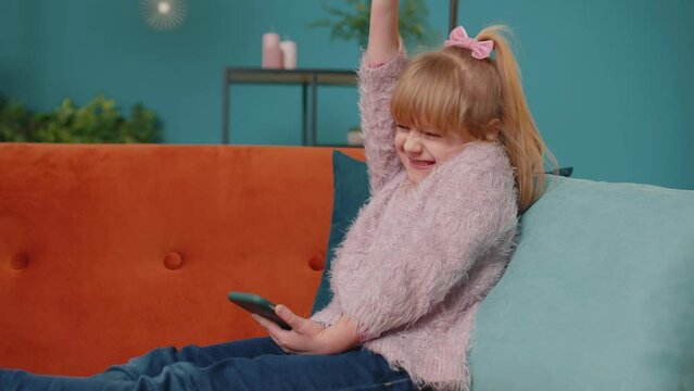 Happy joyful little school kid girl use mobile cell phone browsing say wow yes found out great big win news. Excited child doing winner gesture celebrate holding smartphone lying on sofa alone at home
