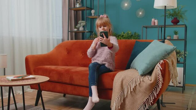 Little teenager toddler school kid girl taking selfie photo with smartphone while sitting on sofa at home. Joyful stylish child make blogger self portrait, holding mobile phone in modern living room