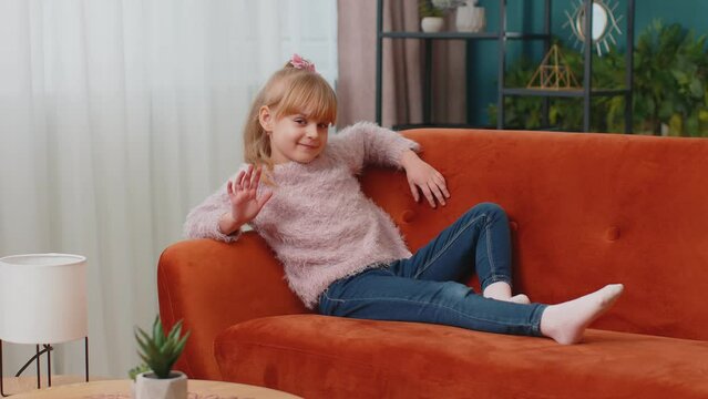 Portrait of relaxed Caucasian little adorable happy child girl lying on sofa at home alone in living room looking at camera, smiling friendly and waving hands gesturing hello or goodbye, welcoming