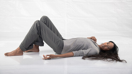 Tall glamorous woman in grey pants and jacket, reclining barefoot in a white studio setting.
