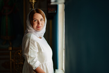 a woman in headscarf and light-colored robes by window in church. 