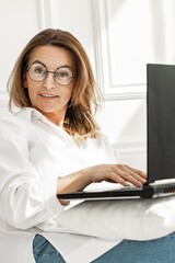 Portrait of an positive middle-aged woman in stylish glasses. Beautiful woman in white cotton shirt sitting on a couch with laptop. Business woman stock photo. White wall background