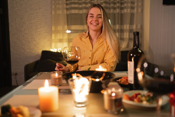 Happy young woman laughing celebrating valentine's day, having dinner and drinking red wine, romantic candlelight dinner, one girl at home table