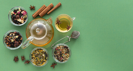 several types of herbal tea mixtures in a glass dish next to a kettle and a cup of hot tea on a green background. View from above. Healing warming aromatic drink. Copy space