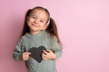 A cute girl with ponytails holds a chalk heart in her hands for recording. Space for text, pink...