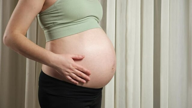 Pregnant woman wearing sports outfit touches stroking bare belly caressing with loving hands standing against window curtain closeup.