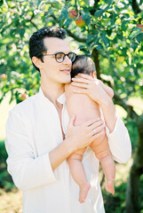 Man in glasses cuddles a newborn to his chest while standing under a tree