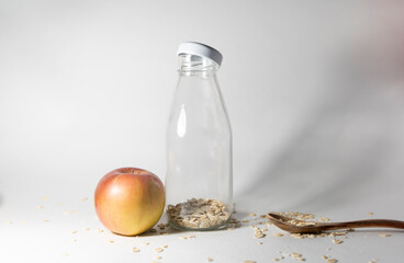 A glass bottle on a white background inside oatmeal. next to it is a red and yellow apple and a wooden spoon. Blank make up. The concept of healthy eating on the way