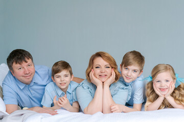 Happy caucasian big family lie in bedroom on bed and smile. Dressed in blue clothes are having fun together. Parents and children two sons and a cute daughter
