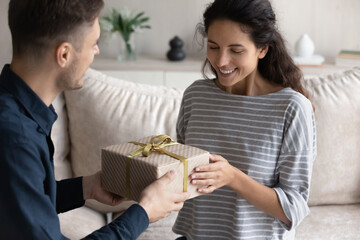 Loving boyfriend congratulating smiling girlfriend, giving gift box, curious beautiful hispanic young woman receiving present, happy young couple celebrating anniversary or birthday at home together