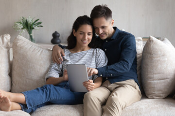 Smiling young couple using tablet together, hugging relaxing sitting on couch, happy woman and man looking at smartphone screen, shopping or chatting online, spending leisure time with gadget at home