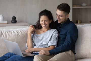 Happy couple using laptop together, relaxing on couch at home, smiling young woman and man holding hands, looking at computer screen, watching movie or funny video, spending leisure time with gadget