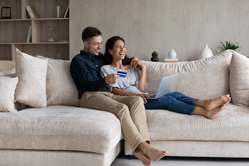 Excited couple paying online by credit card, using laptop, sitting on couch in living room, happy wife and husband shopping, purchasing in internet, making secure payment, browsing banking service