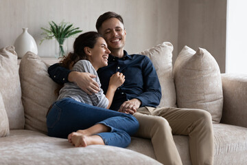 Excited couple hugging, relaxing sitting on couch having fun together, happy beautiful girlfriend and boyfriend spending leisure time lazy weekend at home, talking and laughing at funny joke