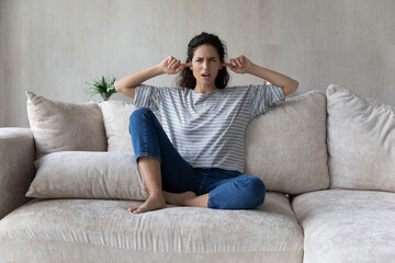 Annoyed unhappy Hispanic woman plugging ears with fingers, sitting on couch at home, noisy disturbing neighbors concept, irritated young female ignoring avoiding loud sound of repairmen - obrazy, fototapety, plakaty