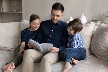 Loving father reading book to little son and daughter, sitting relaxing on cozy couch at home, caring young dad telling to kids funny fairy tale story, spending leisure time weekend together