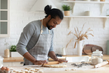 Happy handsome young 30s African American man in apron rolling out dough for homemade pastry, enjoying preparing biscuit cookies in modern light kitchen, cooking hobby activity pastime concept.