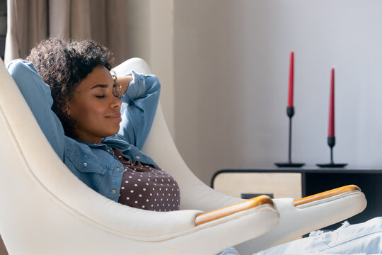 Smiling peaceful young African American woman relaxing in comfortable armchair with folded arms behind head, breathing fresh air, sleeping napping enjoying weekend leisure time, meditating at home.