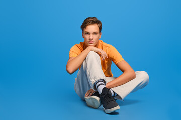 Young man in orange t-shirt and white sweatpants sits on studio floor and propping up his chin over...