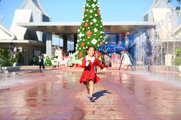 Excited little Asian girl in red dress running to the big christmas tree decorative for the happy new year and merry christmas festival.