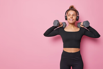 Horizontal shot of sporty healthy woman raises dumbbells trains muscles tries to achieve fitness goals dressed in tracksuit listens music via headphones isolated over pink background blank copy space