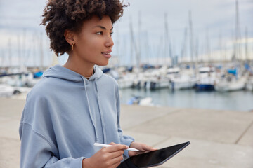 Thoughtful curly haired teenage girl wears casual hoodie poses with modern touch pad and stylus...