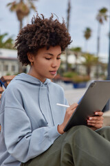 Concentrated curly haired woman wears casual hoodie and trousers makes drawings with stylus pen...