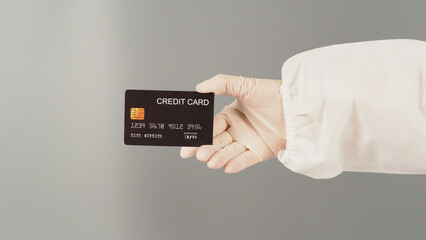 Hand is holding  black credit card on grey background. Hand wears a PPE suit and a white medical glove.