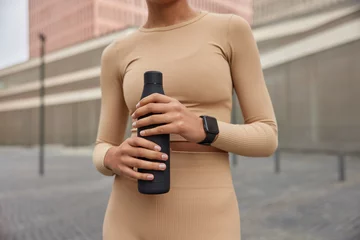 Foto op Plexiglas Cropped shot of unrecognizable woman dressed in sportswear holds sporty bottle of fresh water wears smartwatch feels thirsty after cardio training stands outdoors against blurred background. © Wayhome Studio