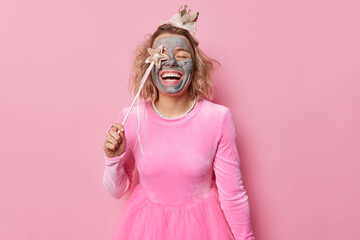 Happy carefree woman covers eye with magic wand entertains children at party applies clay mask undergoes beauty treatments wears crown and dress poses against pink background. Holidays concept - obrazy, fototapety, plakaty