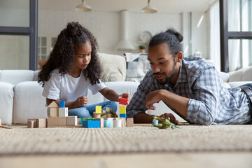 Interested small cute curly African American kid girl constructing building with wooden blocks with caring young father, sitting together on floor carpet in living room, enjoying playing at home.