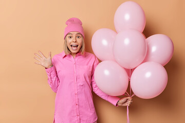 Indoor shot of emotional woman wears pink hat and shirt exclaims loudly holds bunch of helium balloons comes on birthday party isolated over beige background. Festive event and celebration concept