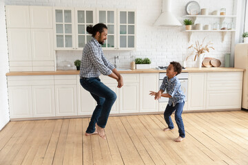 Full length happy caring young African American father dancing barefoot on warm heated wooden floor with joyful laughing adorable little kid boy, listening music having fun together in kitchen.