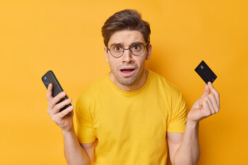 Careless puzzled young man holds mobile phone and credit card feels embarrassed cannot make payment...