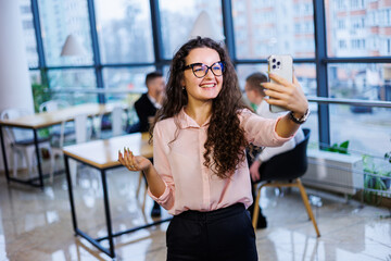Smart happy young business woman, in stylish clothes and glasses, works in the office, talks with a client on a smartphone, gestures, smiles. Employees are working in the background. Selective focus
