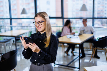 Charming businesswoman with glasses dressed everyday, using a smartphone and talking on the phone, in a modern office.