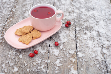 a mug of rosehip tea stands on a wooden table covered with frost. there are cookies on the saucer