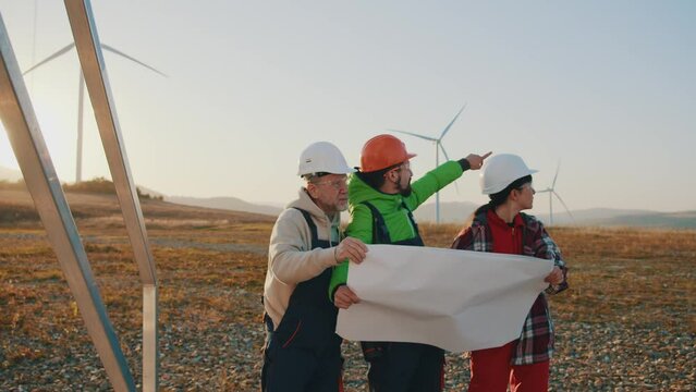 Engineers in hard hat working on ecological station. Groups using a large blueprint on background of wind turbines renewable energetics. Slow motion