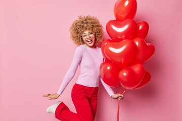 Holiday party and celebration concept. Glad carefree woman celebrates St Valentines Day holds bunch of heart shaped balloons happy to get present from boyfriend wears pink jumper and red trousers