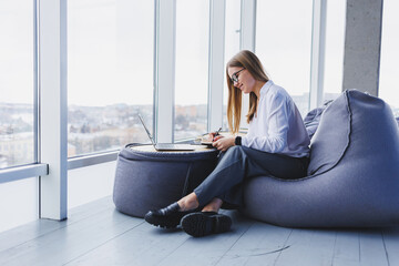 A businesswoman woman looks at the screen sitting in a cafe with a laptop, she makes notes in her...