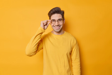 Waist up shot of handsome smiling European adult man keeps hand on rim of spectacles wears casual jumper being in good mood isolated over vivid yellow background. People and emotions concept