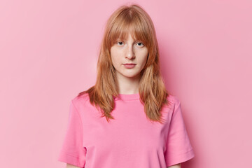 Portrait of serious teenage girl with ginger hair freckles on face looks directly at camera wears casual t shirt isolated on pink background. Pretty redhead schoolgirl has natural beauty poses indoor