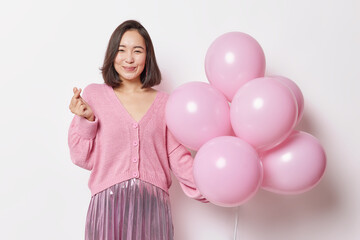Obraz na płótnie Canvas Pleased Asian woman wears fashionable jumper and pleated skirt makes korean like sign holds bunch of inflated balloons celebrates special occasion isolated over white background. Holidays concept