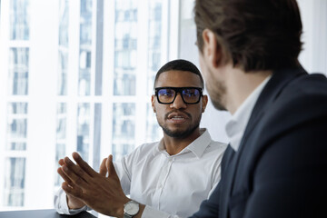 Serious engaged African employee in glasses talking to coworker, business partner, speaking, telling about project. Multiethnic workers, professionals discussing work tasks at workplace